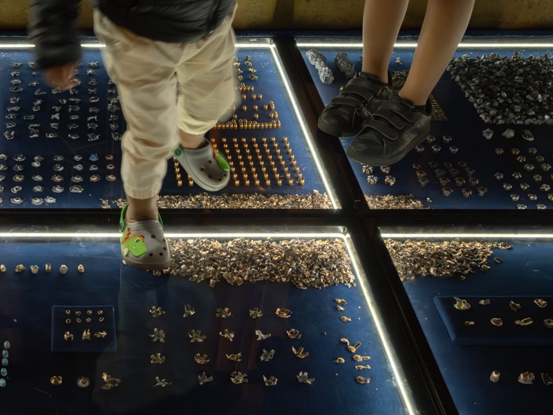 Visitors’ legs and feet over the illuminated modules of the installation, among rows of small objects.
